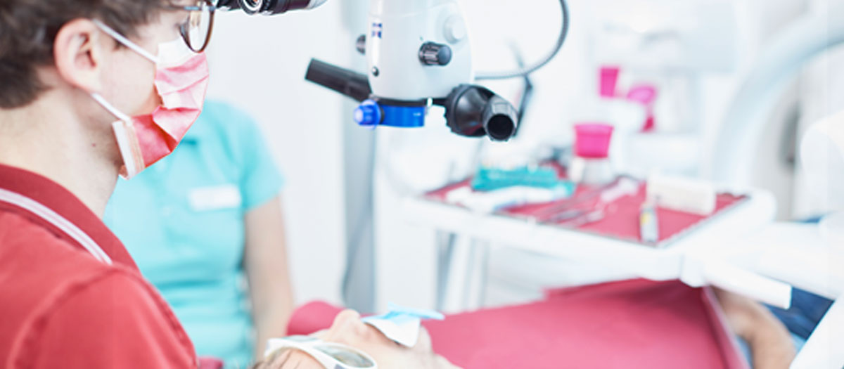 Zahnarzt Doktor Mihlan mit einem Mundschutz behandelt einen Patienten, der auf dem Behandlungsstuhl liegt. Der Patient trägt eine Schutzbrille.